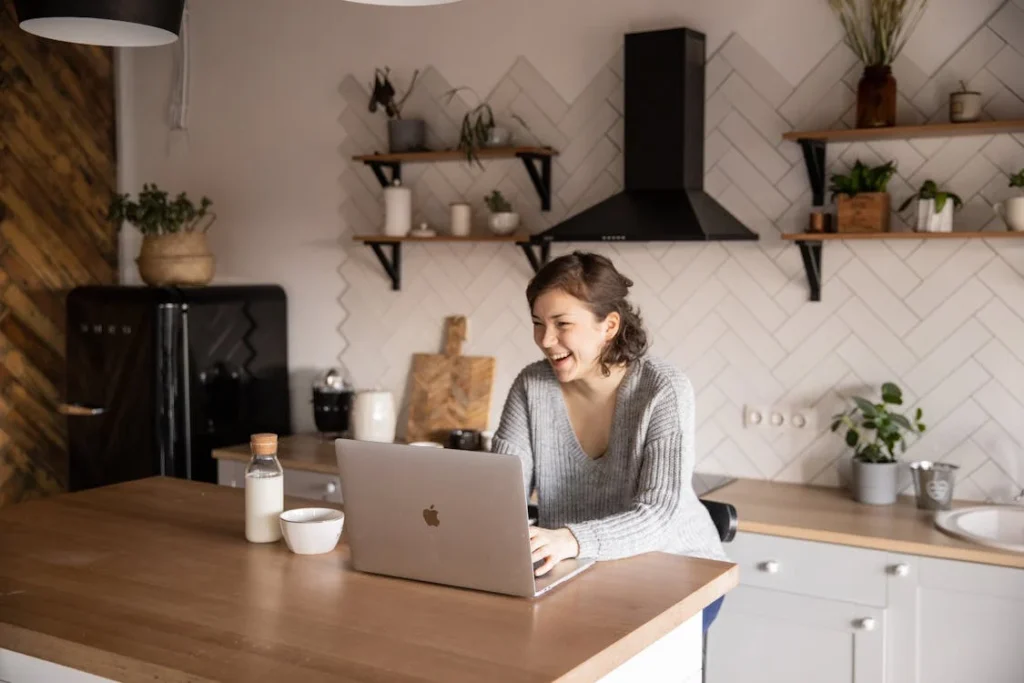 Mujer sonriente en su cocina mientras asiste a una sesión de psicoterapia online en su portátil, disfrutando de la comodidad de su hogar.