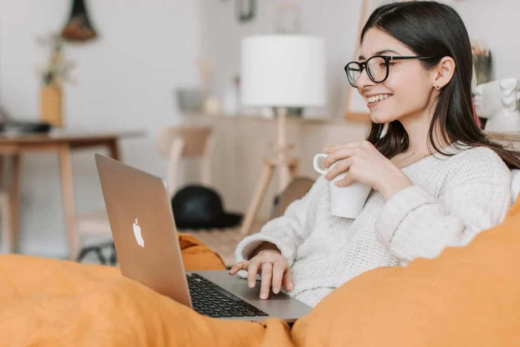Joven con gafas y una taza en la mano, participando en una sesión de terapia online desde su cama, sintiéndose relajada y en un espacio seguro.