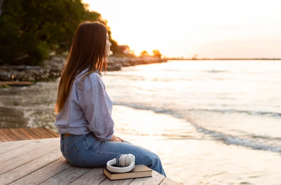 Mujer sentada en una plataforma de madera frente al mar durante el atardecer, con auriculares y un libro sobre la madera, reflexionando y disfrutando de la tranquilidad y equilibrio de la armonización energética