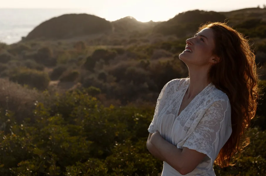 Una mujer con un vestido blanco de pie en un paisaje, mirando hacia el cielo, vibrando en alta frecuencia, iluminada por la suave luz del atardecer.